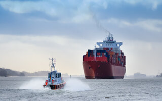 Ein kleines Lotsenboot vor einem riesigen Containerschiff auf dem Fluss Elbe, Deutschland / A small pilot boat in front of a huge container ship on the river Elbe, Germany