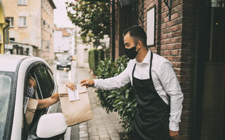 Waiter wearing protective face mask is giving disposable package with food to pretty female driver outside a restaurant