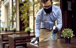 Vorbereitung für die Kunden nach wiedereröffnung / Waiter wearing protective face mask while disinfecting tables at outdoor cafe